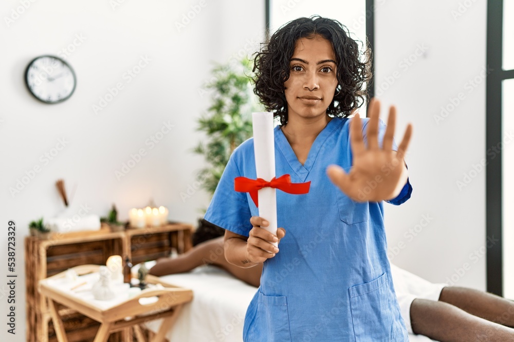 Young therapist woman holding diploma at spa wellness center with open hand doing stop sign with serious and confident expression, defense gesture Young therapist woman holding diploma at spa wellness center with open hand doing stop sign with serious and confident expression, defense gesture