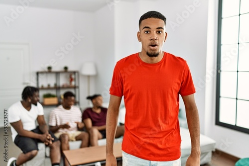Young african man at home with friends sitting on the sofa at home scared and amazed with open mouth for surprise, disbelief face