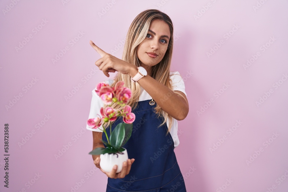 Young blonde woman wearing gardener apron holding plant pointing with hand finger to the side showing advertisement, serious and calm face
