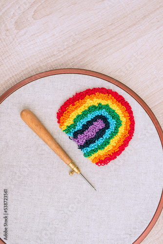 Top view of embroidery hoop with a bulky rainbow, made in a technique of punch needle and a needle below, on white wooden surface.