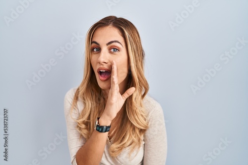 Young blonde woman standing over isolated background hand on mouth telling secret rumor, whispering malicious talk conversation