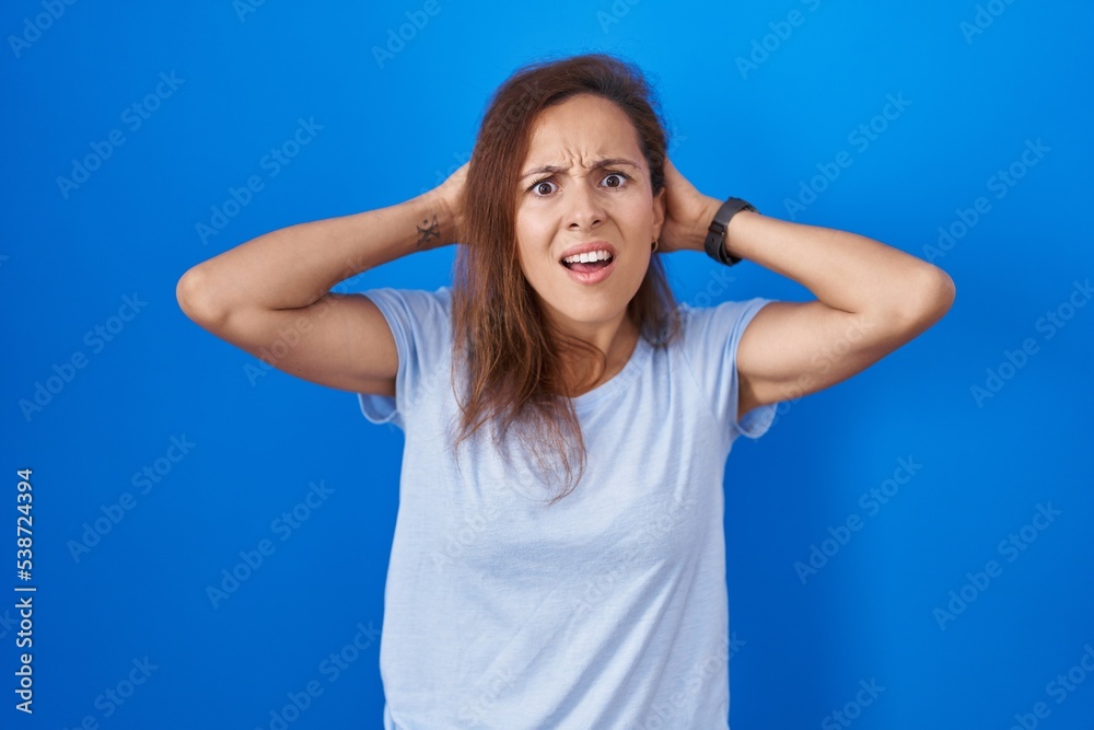 Fototapeta premium Brunette woman standing over blue background crazy and scared with hands on head, afraid and surprised of shock with open mouth