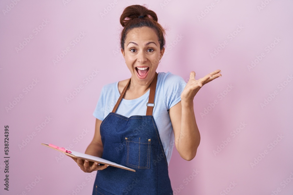 Brunette woman wearing professional waitress apron holding clipboard celebrating victory with happy smile and winner expression with raised hands Brunette woman wearing professional waitress apron holding clipboard celebrating victory with happy smile and winner expression with raised hands