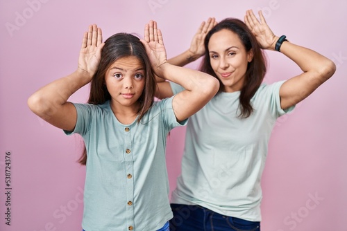 Young mother and daughter standing over pink background doing bunny ears gesture with hands palms looking cynical and skeptical. easter rabbit concept.