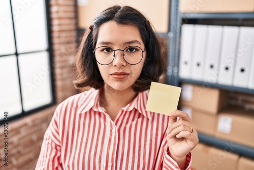 Young hispanic woman holding paper reminder at the office thinking attitude and sober expression looking self confident