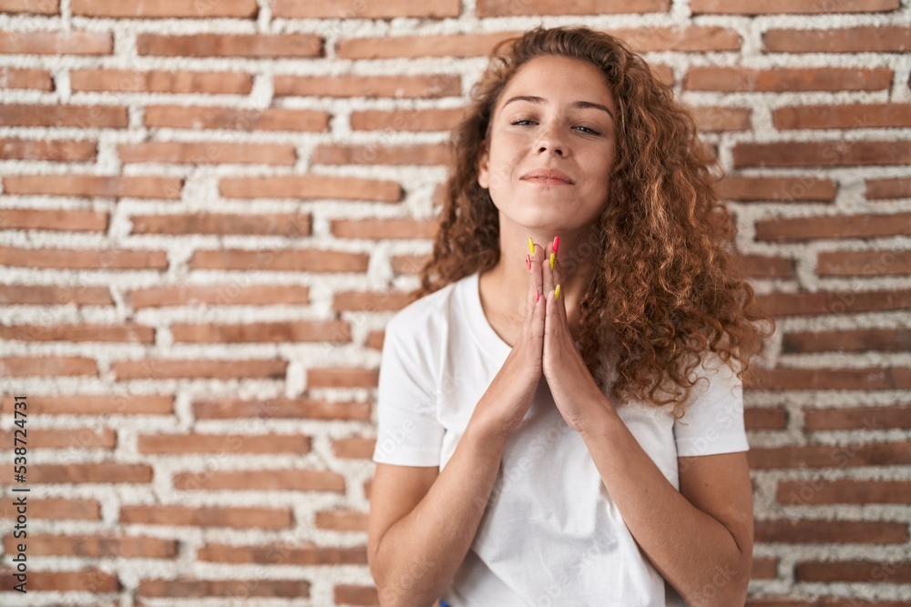 Young caucasian woman standing over bricks wall background praying with hands together asking for forgiveness smiling confident.