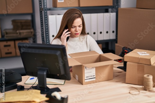 Young blonde woman ecommerce business worker unboxing package and talking on the smartphone at office