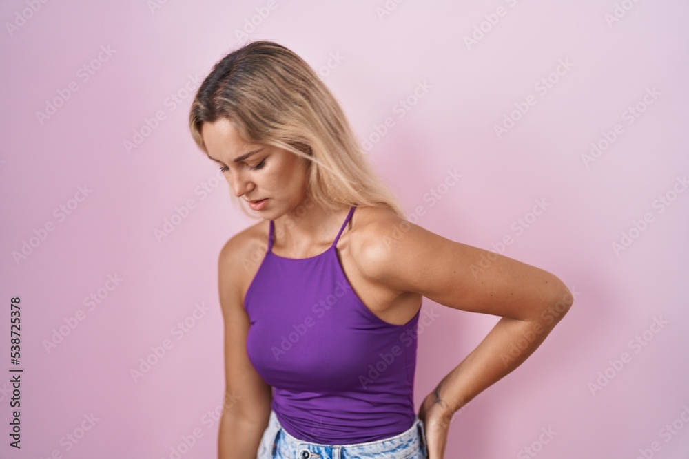 Young blonde woman standing over pink background suffering of backache, touching back with hand, muscular pain Young blonde woman standing over pink background suffering of backache, touching back with hand, muscular pain