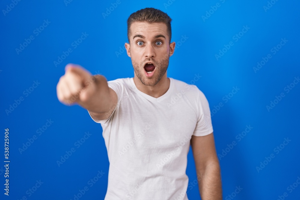 Naklejka premium Young caucasian man standing over blue background pointing with finger surprised ahead, open mouth amazed expression, something on the front
