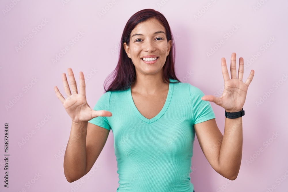 Young hispanic woman standing over pink background showing and pointing up with fingers number ten while smiling confident and happy.