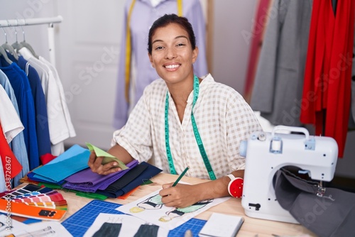 Young beautiful hispanic woman tailor drawing clothing design holding cloth at tailor shop