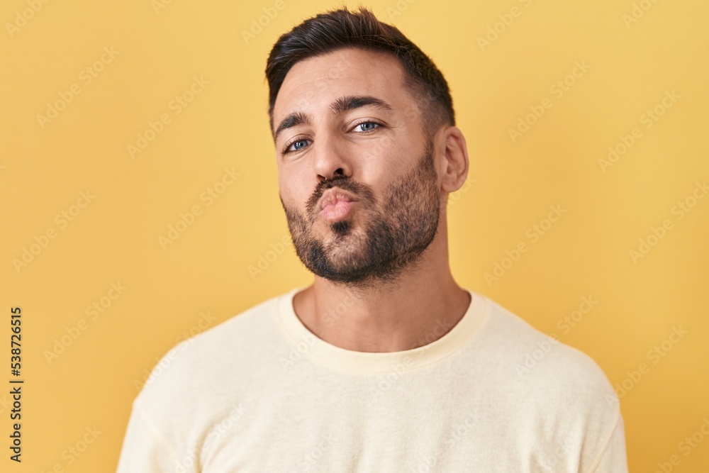 Handsome hispanic man standing over yellow background looking at the camera blowing a kiss on air being lovely and sexy. love expression. Handsome hispanic man standing over yellow background looking at the camera blowing a kiss on air being lovely and sexy. love expression.
