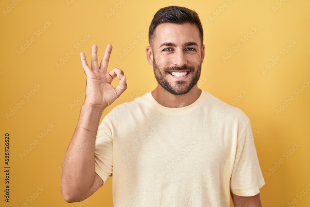 Handsome hispanic man standing over yellow background smiling positive doing ok sign with hand and fingers. successful expression. Handsome hispanic man standing over yellow background smiling positive doing ok sign with hand and fingers. successful expression.