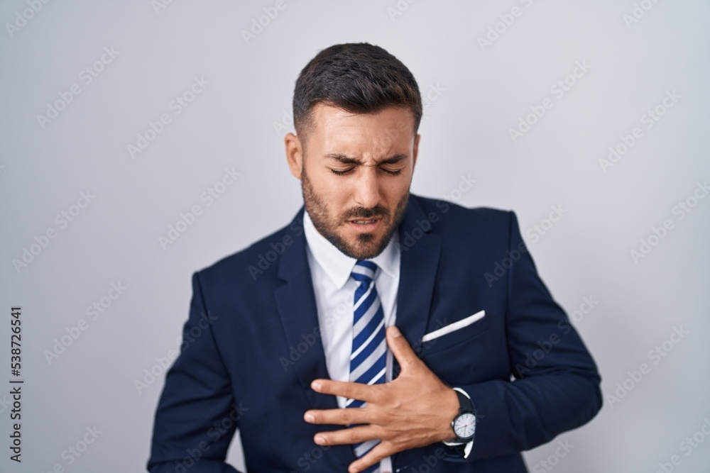 Handsome hispanic man wearing suit and tie with hand on stomach because indigestion, painful illness feeling unwell. ache concept. Handsome hispanic man wearing suit and tie with hand on stomach because indigestion, painful illness feeling unwell. ache concept.