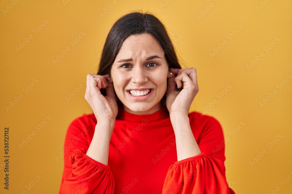Young hispanic woman standing over yellow background covering ears with fingers with annoyed expression for the noise of loud music. deaf concept. Young hispanic woman standing over yellow background covering ears with fingers with annoyed expression for the noise of loud music. deaf concept.
