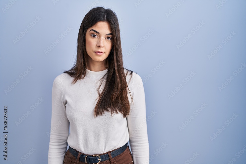 Obraz premium Young brunette woman standing over blue background relaxed with serious expression on face. simple and natural looking at the camera.