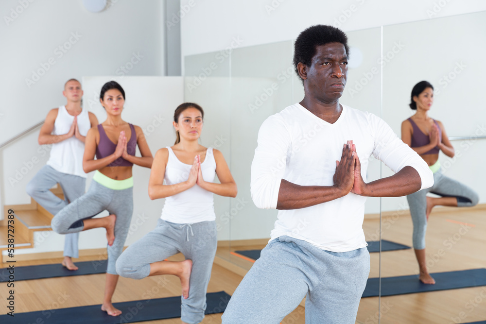 Focused aframerican man standing on one leg in Eka Pada Pranamasana pose during group yoga training in gym