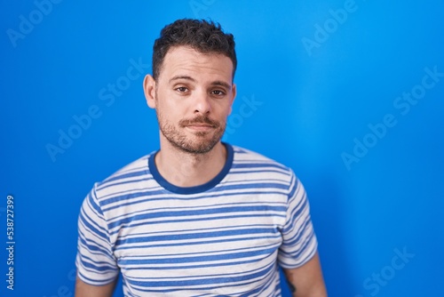 Young hispanic man standing over blue background looking sleepy and tired, exhausted for fatigue and hangover, lazy eyes in the morning.