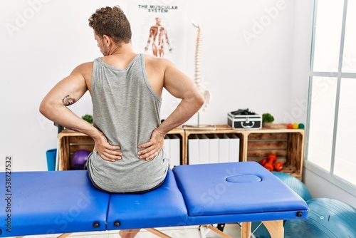 Young hispanic man touching injury back sitting on massage board at clinic