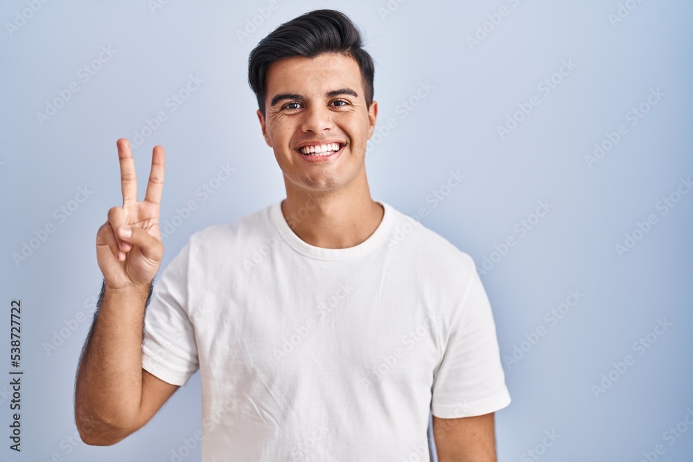 Hispanic man standing over blue background showing and pointing up with fingers number two while smiling confident and happy. Hispanic man standing over blue background showing and pointing up with fingers number two while smiling confident and happy.