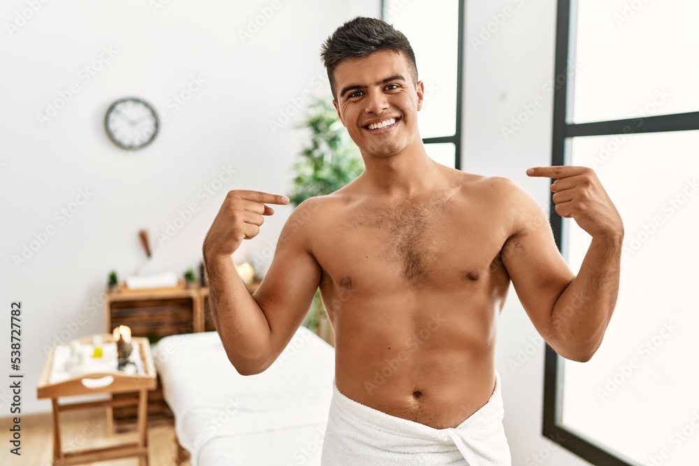 Young hispanic man standing shirtless at spa center looking confident with smile on face, pointing oneself with fingers proud and happy. Young hispanic man standing shirtless at spa center looking confident with smile on face, pointing oneself with fingers proud and happy.