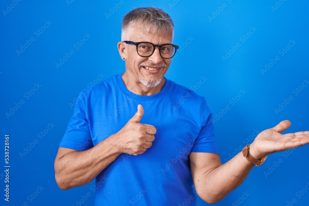 Hispanic man with grey hair standing over blue background showing palm hand and doing ok gesture with thumbs up, smiling happy and cheerful Hispanic man with grey hair standing over blue background showing palm hand and doing ok gesture with thumbs up, smiling happy and cheerful