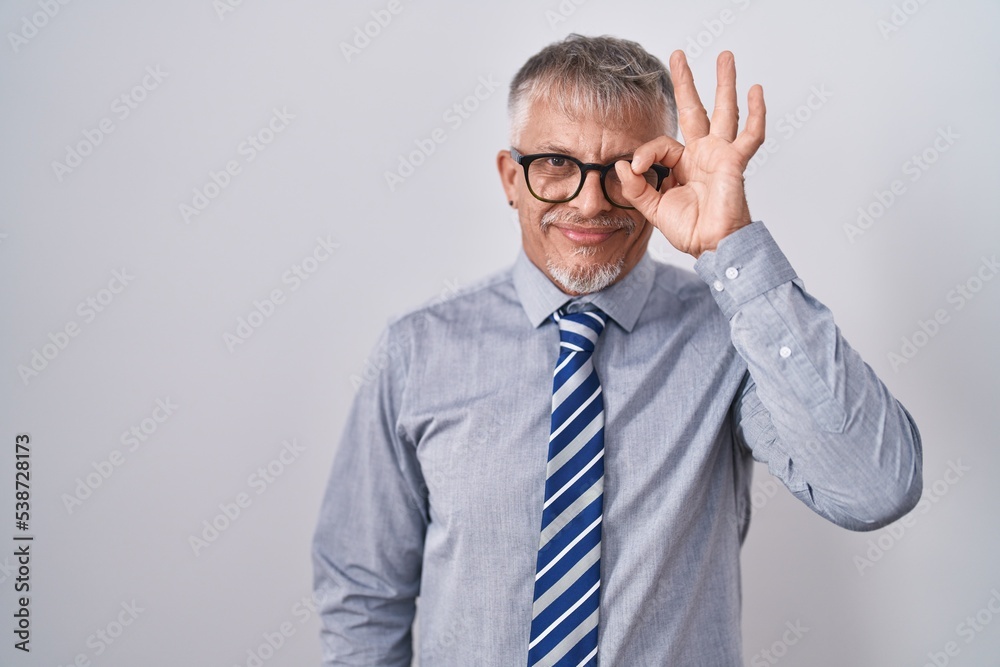 Hispanic business man with grey hair wearing glasses doing ok gesture with hand smiling, eye looking through fingers with happy face. Hispanic business man with grey hair wearing glasses doing ok gesture with hand smiling, eye looking through fingers with happy face.