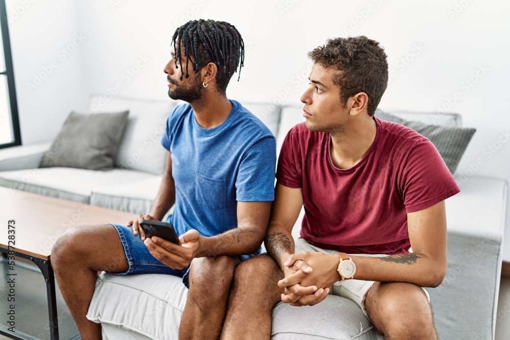 Fototapeta premium Young hispanic men using smartphone sitting on the sofa at home looking to side, relax profile pose with natural face and confident smile.