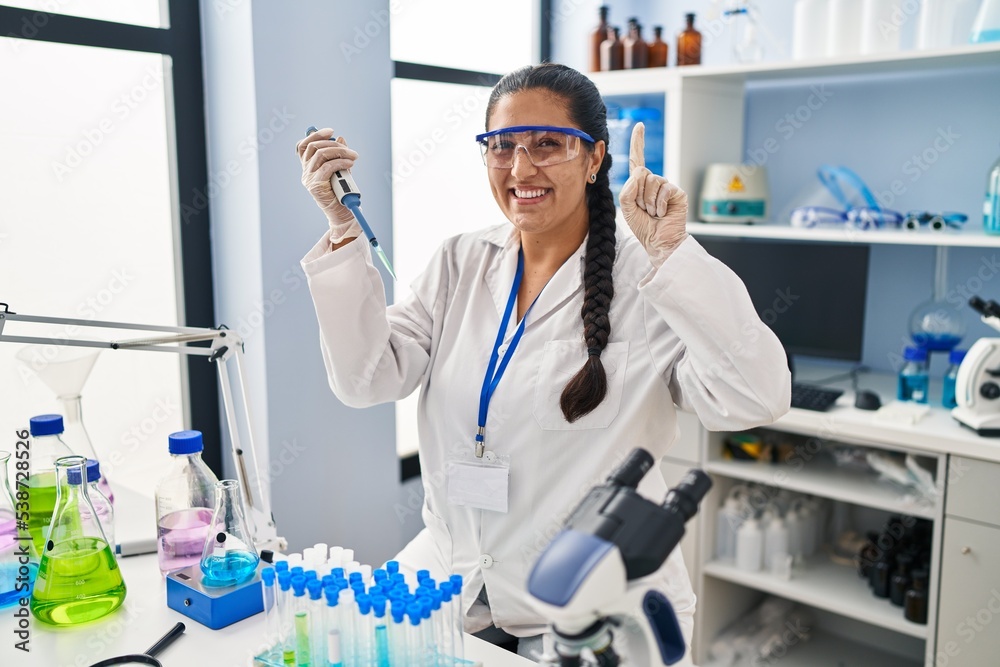 Young hispanic woman working at scientist laboratory smiling with an idea or question pointing finger with happy face, number one Young hispanic woman working at scientist laboratory smiling with an idea or question pointing finger with happy face, number one