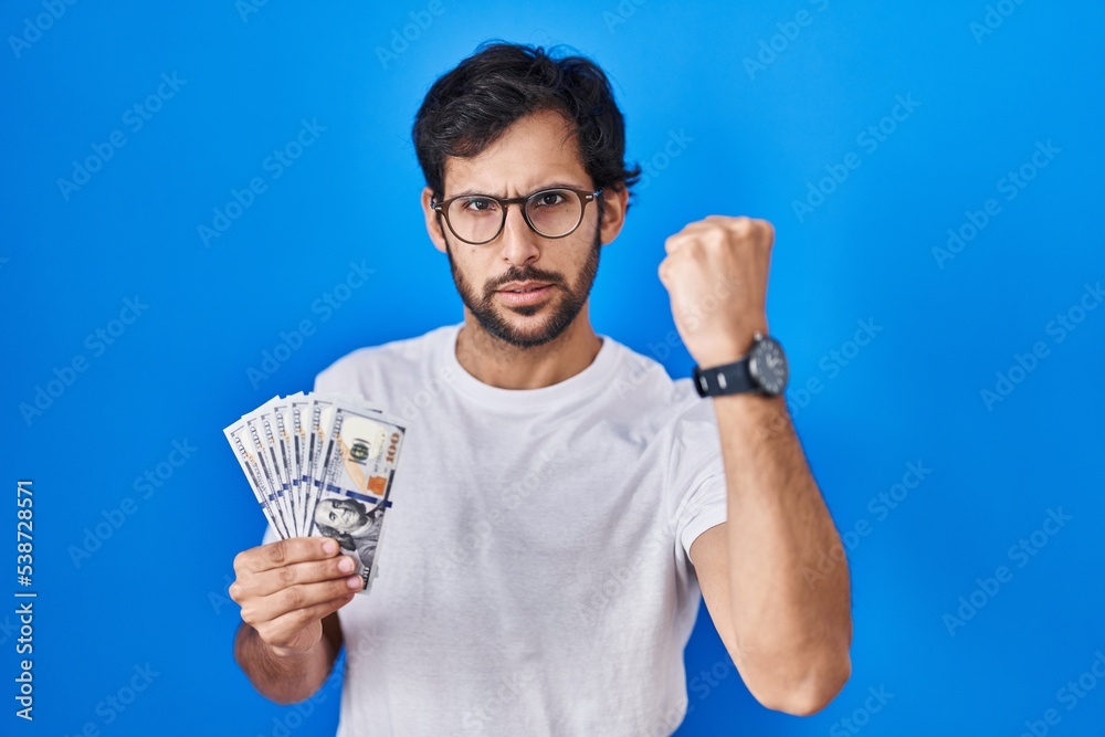Handsome latin man holding dollars banknotes annoyed and frustrated shouting with anger, yelling crazy with anger and hand raised Handsome latin man holding dollars banknotes annoyed and frustrated shouting with anger, yelling crazy with anger and hand raised