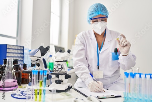 Middle age woman wearing scientist unifor and medical mask holding vaccine write on clipboard at laboratory