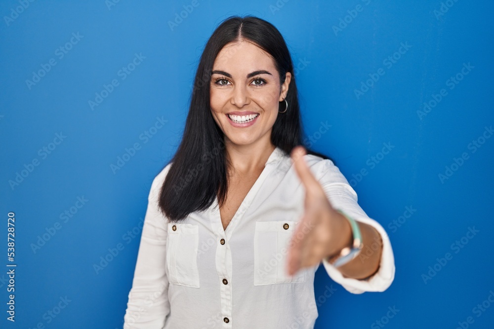 Young hispanic woman standing over blue background smiling friendly offering handshake as greeting and welcoming. successful business. Young hispanic woman standing over blue background smiling friendly offering handshake as greeting and welcoming. successful business.