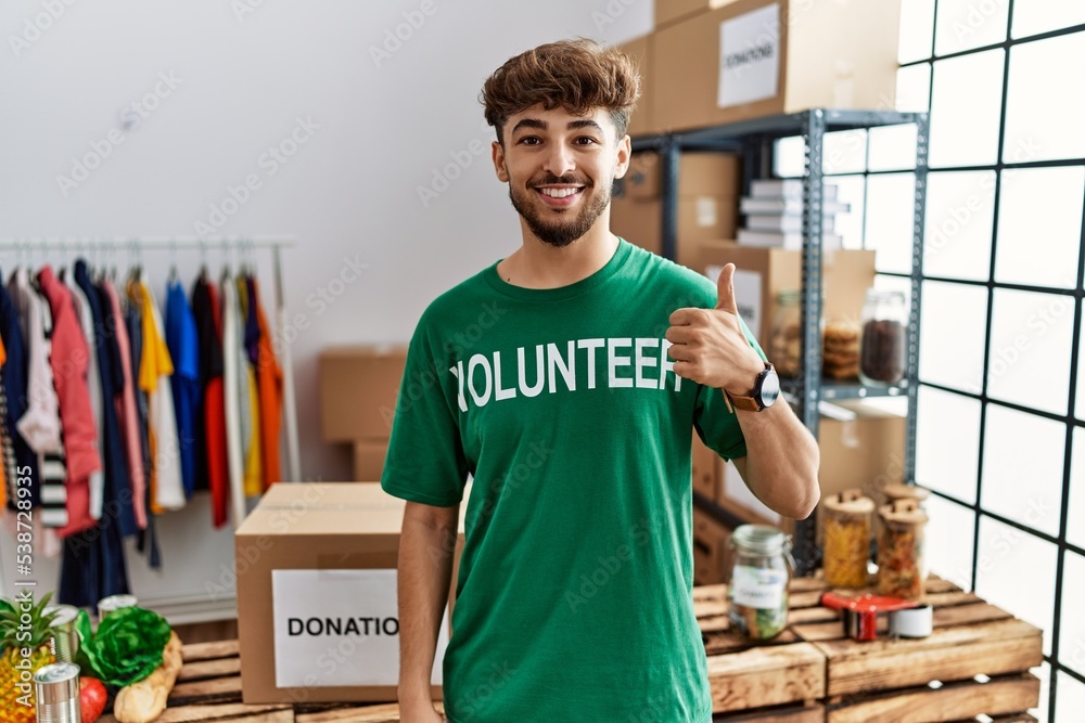 Young arab man wearing volunteer t shirt at donations stand doing happy thumbs up gesture with hand. approving expression looking at the camera showing success. Young arab man wearing volunteer t shirt at donations stand doing happy thumbs up gesture with hand. approving expression looking at the camera showing success.