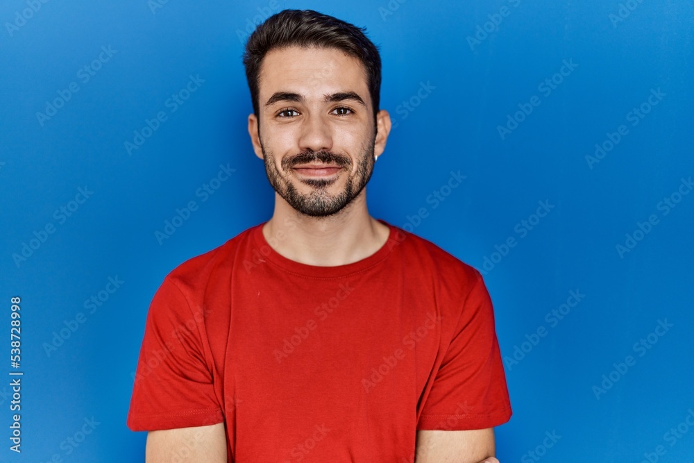Young hispanic man with beard wearing red t shirt over blue background with a happy and cool smile on face. lucky person. Young hispanic man with beard wearing red t shirt over blue background with a happy and cool smile on face. lucky person.