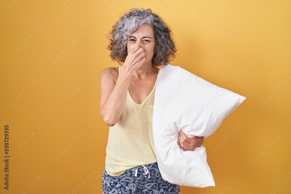 Middle age woman with grey hair wearing pijama hugging pillow smelling something stinky and