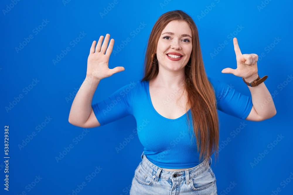 Redhead woman standing over blue background showing and pointing up with fingers number seven while smiling confident and happy. Redhead woman standing over blue background showing and pointing up with fingers number seven while smiling confident and happy.