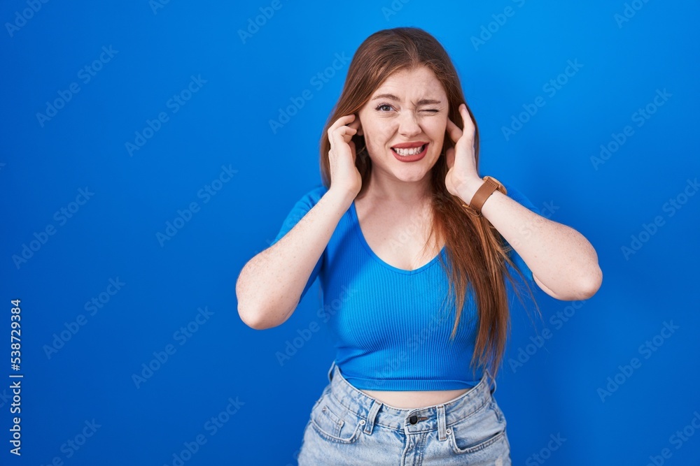Redhead woman standing over blue background covering ears with fingers with annoyed expression for the noise of loud music. deaf concept. Redhead woman standing over blue background covering ears with fingers with annoyed expression for the noise of loud music. deaf concept.