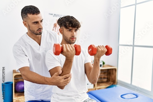 Two hispanic men physiotherapist and patient having rehab session using dumbbells at clinic