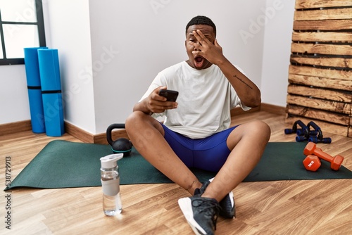 Young african man sitting on training mat at the gym using smartphone peeking in shock covering face and eyes with hand, looking through fingers with embarrassed expression.