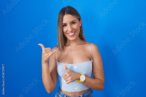 Young woman standing over blue background pointing to the back behind with hand and thumbs up, smiling confident © Krakenimages.com