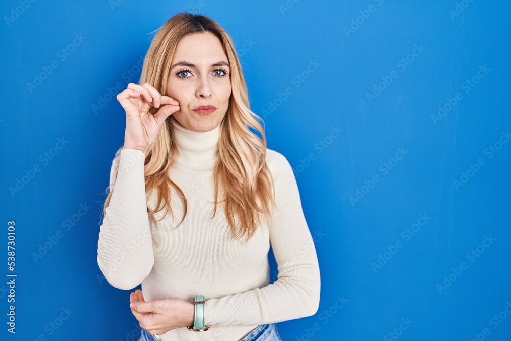 Young caucasian woman standing over blue background mouth and lips shut as zip with fingers. secret and silent, taboo talking Young caucasian woman standing over blue background mouth and lips shut as zip with fingers. secret and silent, taboo talking