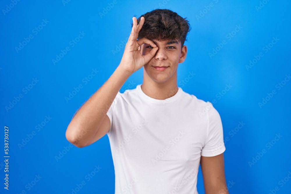 Hispanic teenager standing over blue background doing ok gesture with hand smiling, eye looking through fingers with happy face. Hispanic teenager standing over blue background doing ok gesture with hand smiling, eye looking through fingers with happy face.