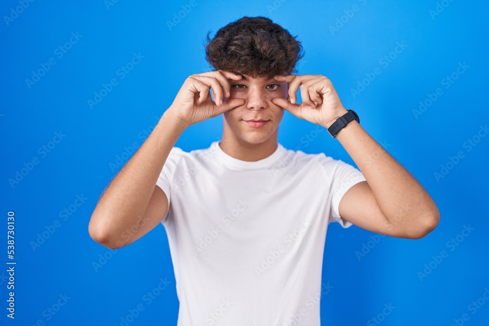 Hispanic teenager standing over blue background trying to open eyes with fingers, sleepy and tired for morning fatigue