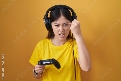Chinese young woman playing video game holding controller angry and mad raising fist frustrated and furious while shouting with anger. rage and aggressive concept.