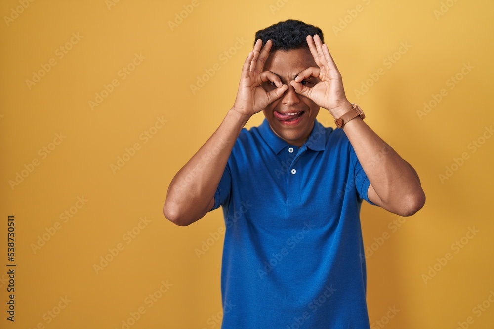Young hispanic man standing over yellow background doing ok gesture like binoculars sticking tongue out, eyes looking through fingers. crazy expression. Young hispanic man standing over yellow background doing ok gesture like binoculars sticking tongue out, eyes looking through fingers. crazy expression.