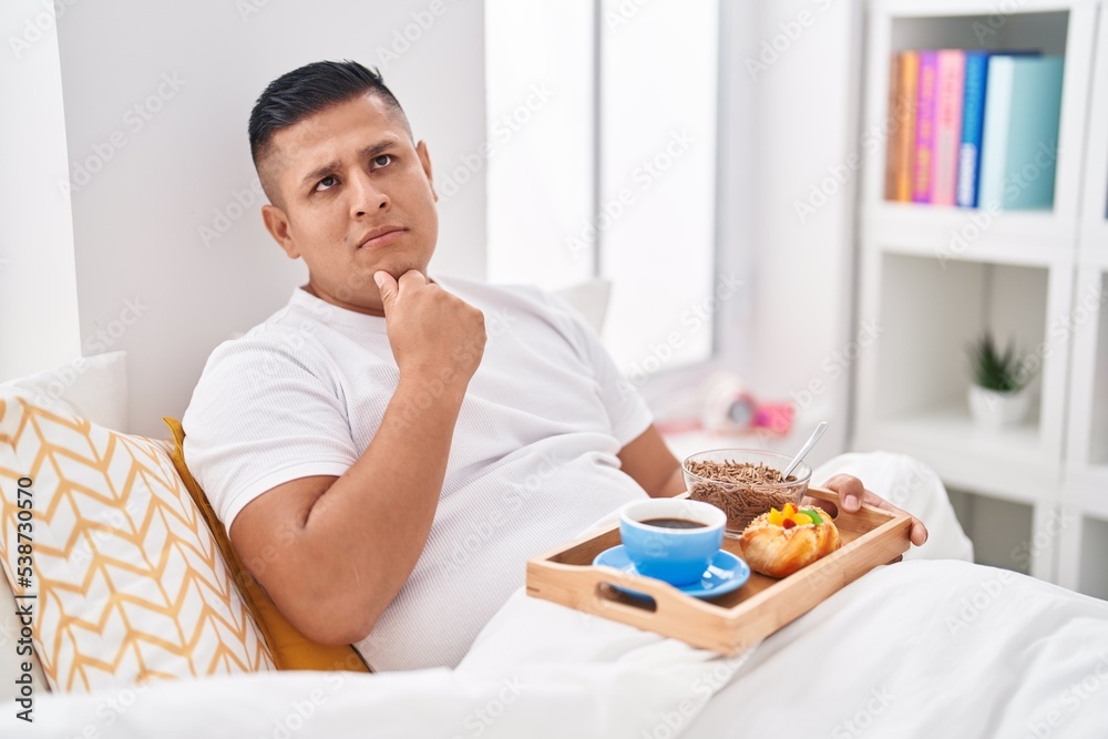 Young hispanic man eating breakfast in the bed serious face thinking about question with hand on chin, thoughtful about confusing idea Young hispanic man eating breakfast in the bed serious face thinking about question with hand on chin, thoughtful about confusing idea