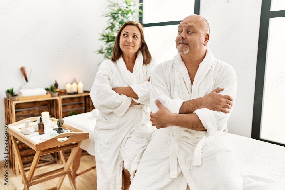 Middle age hispanic couple wearing bathrobe at wellness spa smiling looking to the side and staring away thinking.
