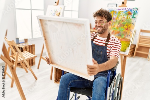 Young hispanic man sitting on wheelchair holding canvas at art studio