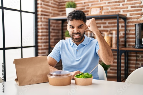 Hispanic man with beard eating delivery salad annoyed and frustrated shouting with anger, yelling crazy with anger and hand raised