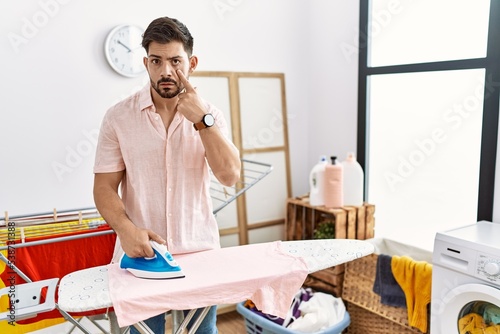 Young man with beard ironing clothes at home pointing to the eye watching you gesture  suspicious expression